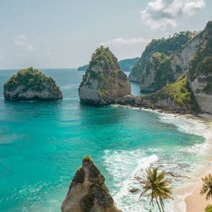 Vue plongeante d'une crique isolée avec de grandes formations rocheuses dans des eaux turquoises, une plage de sable et des falaises abruptes.