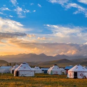 Un campamento de yurtas blancas se asienta en un campo de hierba con montañas al fondo bajo un cielo al atardecer.