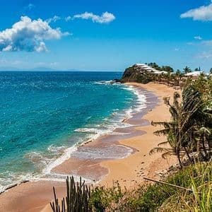 Vista aerea di una spiaggia tropicale sabbiosa con acqua turchese e palme lungo la costa, sotto un cielo azzurro con nuvole.