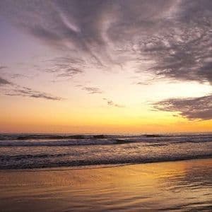 Un tramonto dorato si riflette sulla sabbia bagnata di una spiaggia mentre le onde si infrangono sotto un cielo nuvoloso con sfumature viola.