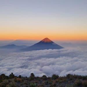 La cima di un vulcano si erge sopra un mare di nuvole, illuminata dalla luce dorata dell'alba, vista da un punto panoramico roccioso.