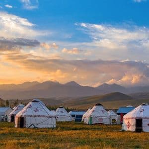 Un accampamento di yurte bianche in un campo erboso con una catena montuosa visibile sullo sfondo sotto un cielo al tramonto.