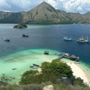 Una vista dall'alto di barche su una baia tropicale con acqua turchese, una spiaggia di sabbia bianca, un molo di legno e colline verdi.