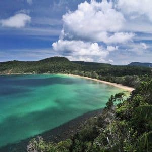 Una vista dall'alto di una cala tropicale con acqua turchese, una spiaggia sabbiosa e una collina boscosa sotto un cielo parzialmente nuvoloso.