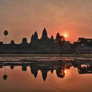 Un antico complesso di templi si staglia contro un cielo arancione all'alba, con il suo riflesso proiettato sull'acqua immobile di un lago.