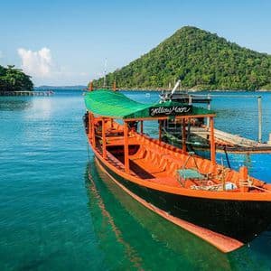 Ein farbenfrohes Longtail-Boot mit grünem Dach liegt in ruhigem, türkisfarbenem Wasser vor einem bewaldeten Hügel.