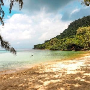 Una spiaggia sabbiosa con l'ombra degli alberi che conduce ad acque calme e limpide, dove due persone nuotano vicino a una collina boscosa.