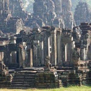 Antiche rovine in pietra di un tempio, con pilastri e torri adornate da grandi volti scolpiti, immerse in una foresta nebbiosa.