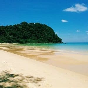 Ein breiter Sandstrand trifft auf ruhiges, türkisfarbenes Wasser neben einem üppigen, bewaldeten Hügel unter einem klaren blauen Himmel.