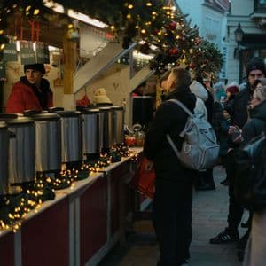 Auf einem Weihnachtsmarkt im Freien stehen Menschen in Wintermänteln an einem Stand Schlange, um heiße Getränke aus großen Edelstahlspendern zu bekommen.