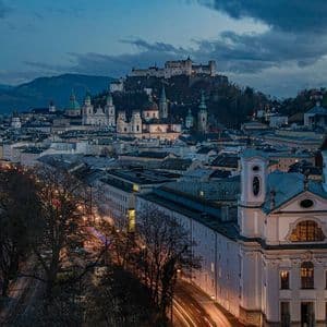 Beleuchtete Stadtansicht bei Dämmerung mit Festung auf Hügel, historischen Gebäuden und entfernten Bergen unter bewölktem Himmel.