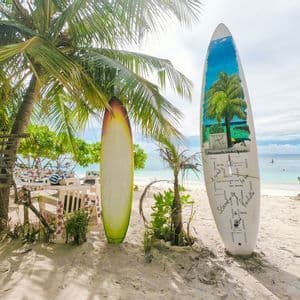 Due tavole da surf si ergono dritte nella sabbia sotto le palme su una spiaggia tropicale, con un'area salotto e l'oceano sullo sfondo.