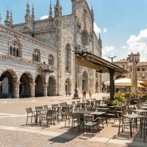 Un caffè all'aperto con tavoli vuoti si trova in una piazza soleggiata e pavimentata di fronte a una grande cattedrale in pietra a strisce.