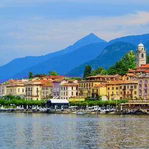 Una colorata cittadina in riva al lago con un campanile è incastonata ai piedi di grandi montagne alberate sotto un cielo azzurro.