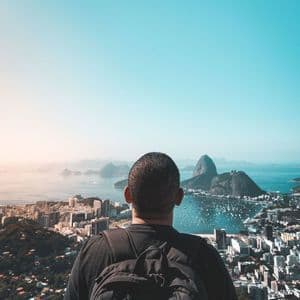 A person wearing a backpack looks out over a panoramic view of a coastal city and a bay with mountains.
