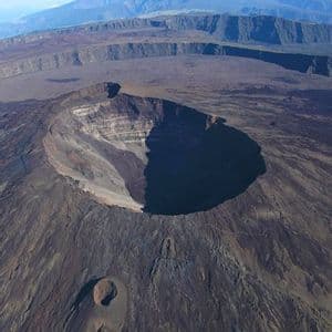 Vue aérienne d'un grand cratère volcanique dans un vaste paysage rocheux avec des montagnes lointaines sous un ciel bleu.