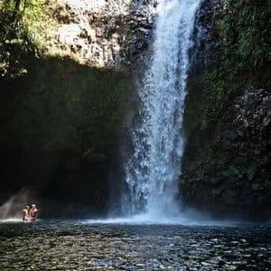 Un voyage de groupe WeRoad se baignant dans l'eau au pied d'une grande cascade qui dévale une falaise rocheuse escarpée.