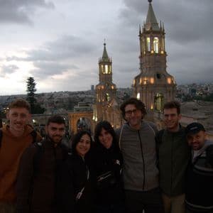 Un viaggio di gruppo WeRoad di sette persone in posa su un punto panoramico con due torri di cattedrale illuminate e uno skyline della città al tramonto.