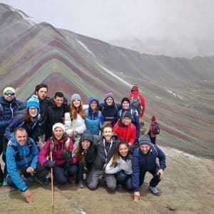 Eine WeRoad-Gruppenreise posiert für ein Foto auf einem Berggipfel mit farbenfrohen, geschichteten Felsformationen im Hintergrund.