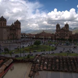 Una vista dall'alto di una vivace piazza storica con due grandi cattedrali in pietra, un parco centrale e le montagne sullo sfondo.