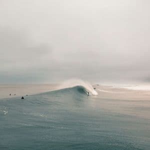 Un surfista cavalca una grande onda tubante nell'oceano, mentre altri surfisti attendono sotto un cielo nuvoloso.