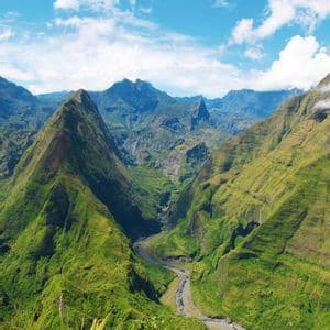 Une rivière sinueuse coule à travers une vallée profonde, entourée de montagnes escarpées et verdoyantes sous un ciel bleu parsemé de nuages.