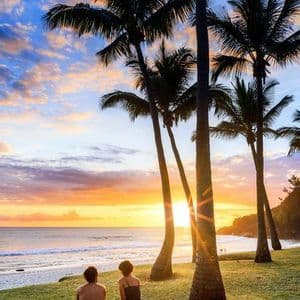 Deux personnes assises sur une couverture jaune sur un rivage herbeux, observant le coucher de soleil sur l'océan derrière de grands palmiers.