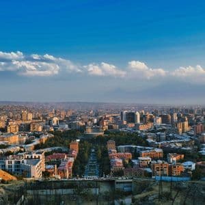 Veduta aerea di una città con un viale centrale, edifici e montagne sullo sfondo sotto un cielo blu e nuvoloso.