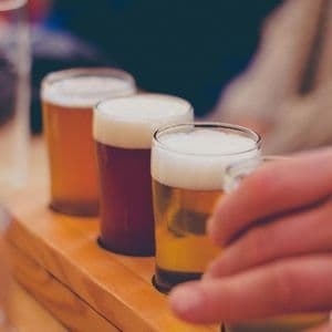 Gros plan sur une main tendant vers un verre lors d'une dégustation de bières, avec quatre bières différentes sur une planche en bois.