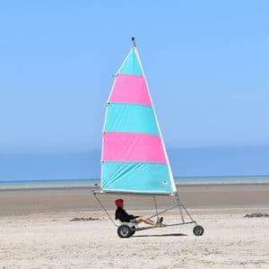 Une personne dans un char à voile avec une voile rose et turquoise est assise sur une large plage de sable près de l'océan sous un ciel bleu clair.