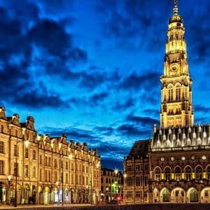 Une haute tour de l'horloge illuminée et des bâtiments historiques bordent une place de ville pavée sous un ciel nocturne nuageux.