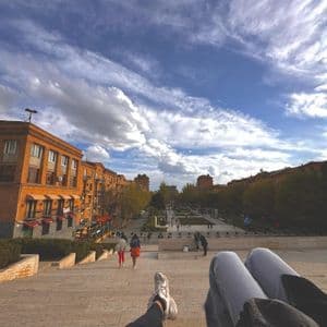 Una vista in prima persona di due persone sedute su un'ampia scalinata in pietra che si affaccia su una piazza cittadina con persone che camminano sotto.