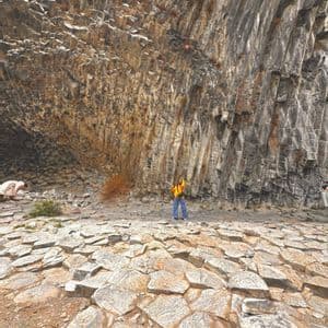 Una donna in gilet arancione si trova su formazioni rocciose esagonali davanti a una parete curva di colonne di basalto.