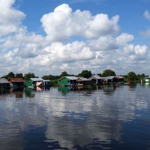 Un village flottant avec des maisons colorées sur une rivière calme, reflétant le ciel bleu et les nuages blancs.