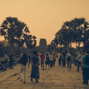 Un voyage de groupe WeRoad emprunte une chaussée de pierre vers d'anciens temples entourés de palmiers sous un ciel doré.