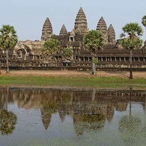 Un ancien temple de pierre avec plusieurs tours et des palmiers se reflète dans une étendue d'eau calme au premier plan.