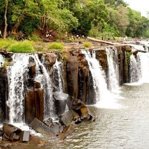 Une large cascade dévale sur des rochers sombres dans une rivière, avec un groupe de voyage WeRoad réuni sur la berge boisée en amont.