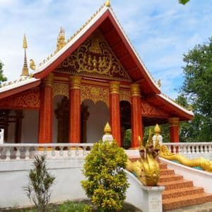 Un temple bouddhiste orné de rouge et d'or, avec des statues de naga dorées le long de son escalier, se dressant sur fond d'arbres et de ciel bleu.