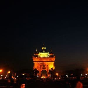 Un grand monument orné illuminé de lumières dorées sur un ciel nocturne sombre, avec des réverbères et des arbres en contrebas.