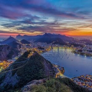 A panoramic view of an illuminated coastal city nestled between mountains and a bay under a colorful sunset sky.