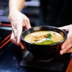 A person serves a bowl of ramen soup with toppings, placing it on a black tray.