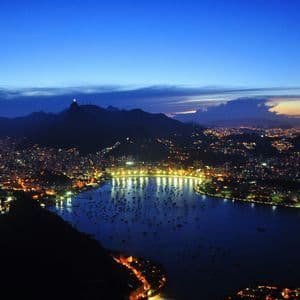 An aerial view of a coastal city illuminated at dusk, with a bay full of boats and mountains silhouetted in the background.