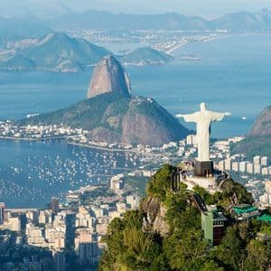 The Christ the Redeemer statue stands on a mountain peak overlooking a coastal city, Sugarloaf Mountain, and a bay filled with boats.