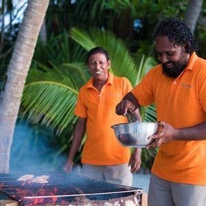 Due uomini con polo arancioni abbinate sorridono mentre preparano cibo su una griglia a carbone all'aperto, con palme sullo sfondo.