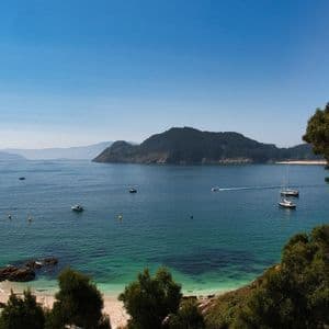 Una vista aérea de una bahía soleada con agua turquesa, una pequeña playa de arena y varios barcos navegando cerca de una isla boscosa.