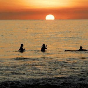 Tres personas de un viaje en grupo de WeRoad silueteadas en el océano durante un atardecer anaranjado, una de ellas en una tabla de surf.