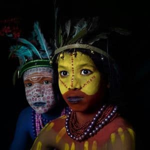 Primer plano de dos jóvenes con elaborada pintura facial tradicional y tocados de plumas sobre fondo negro.