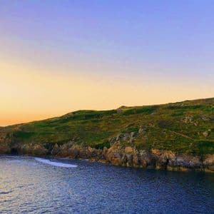 Un faro de piedra se asienta en una colina cubierta de hierba con vistas a una costa rocosa y un mar en calma al atardecer.