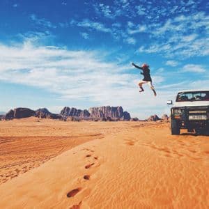 A person jumps in the air on an orange sand dune in the desert, next to a white pickup truck, with mountains in the background.