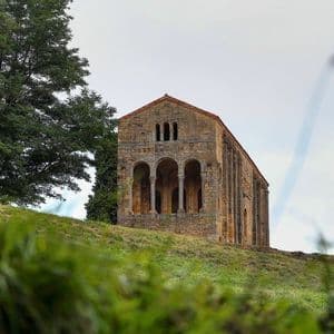 Una antigua iglesia de piedra con fachada arqueada se erige en una colina de hierba junto a un gran árbol bajo un cielo nublado.